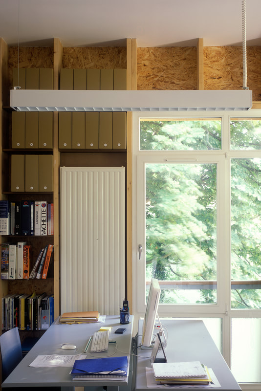 Intérieur de l'atelier : bureau, rangements en bois et grande baie vitrée sur la végétation