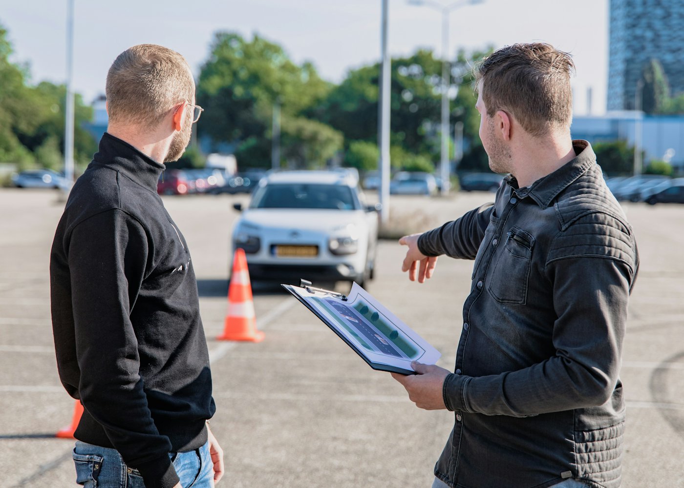 Un enseignant de la conduite explique un exercice à un élève devant la voiture-école, sur une piste balisée de cônes