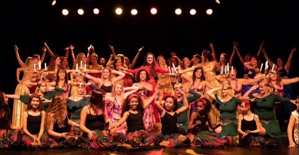 Sur scène, une quarantaine de danseuses en costumes colorés et dorés posent bras levés autour d'Hestia en rouge, au centre