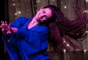 Hestia souriante en tenue khaleegi bleue, cheveux en mouvement, dansant devant un rideau lumineux