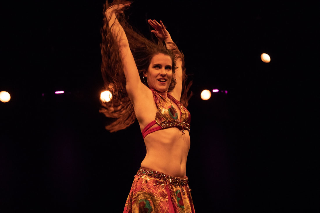 Hestia en costume de danse orientale rouge et or, bras levés, sur scène sous les projecteurs