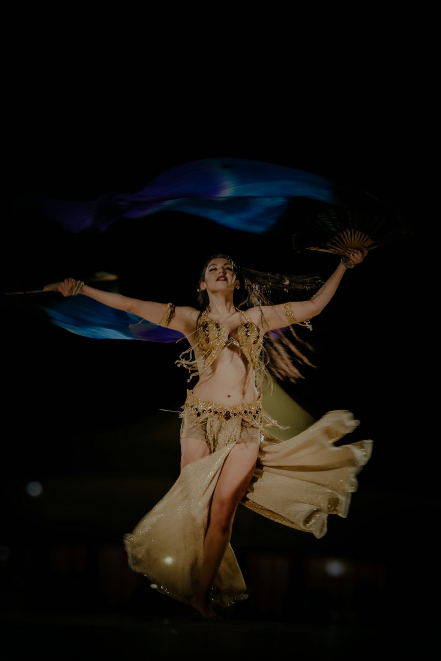 Danseuse orientale en costume doré faisant voler un grand voile bleu sur une scène sombre