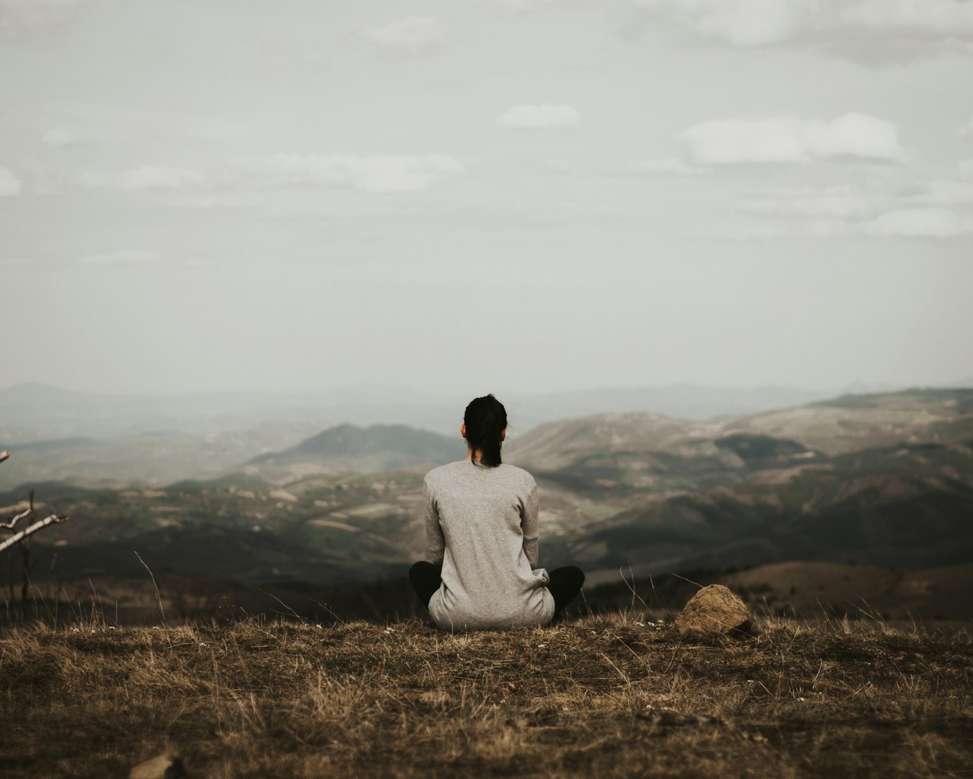 Une personne assise dans l’herbe, de dos, contemplant un paysage de collines brumeuses.